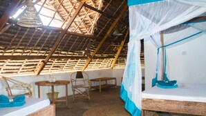 Una habitación con techo de paja, una cama con mosquitero, una silla de mimbre, una mesita y un tapete tejido en el suelo.