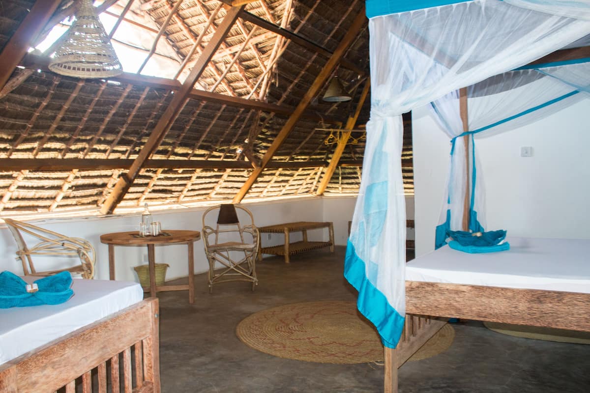A room with a thatched roof, a bed with a mosquito net, a wicker chair, a small table, and a woven mat on the floor.