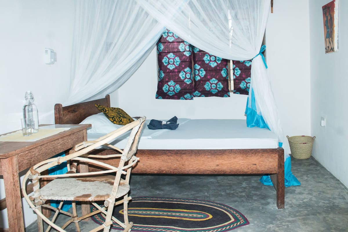 A single bed with a canopy, a wooden chair, a small table with a glass bottle, and a woven basket on the floor.