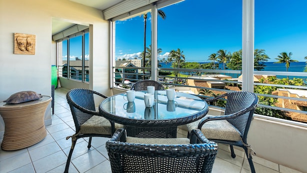 Un balcon avec une table en verre et des chaises en osier, offrant une vue sur la côte, avec des palmiers et des bâtiments.