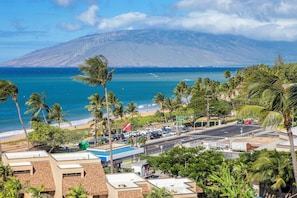 A coastal area with a view of the ocean and a mountain. There are palm trees, a beach, and parked cars.