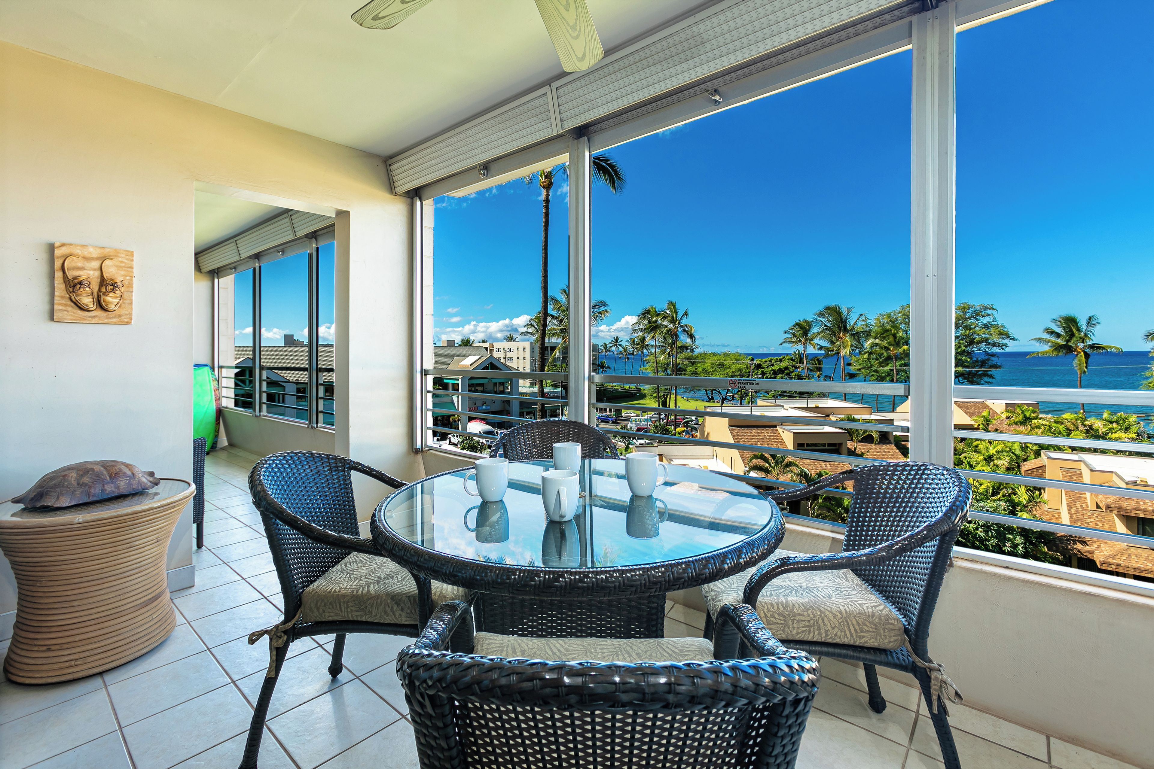 A balcony with a glass table and chairs, overlooking a coastal view with palm trees and buildings.