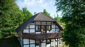 Un chalet traditionnel doté d’un balcon, de grandes fenêtres et d’un toit en pente, entouré d’arbres.