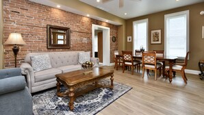 A living room with a brick wall, a ceiling fan, a dining area with wooden furniture, and a large rug.