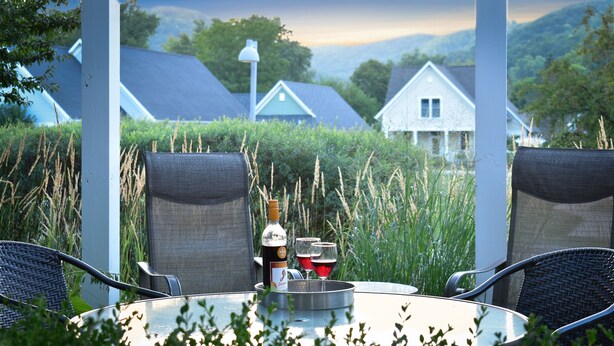 A round glass table with two wicker chairs, a bottle, and two glasses of red wine on a metal tray.