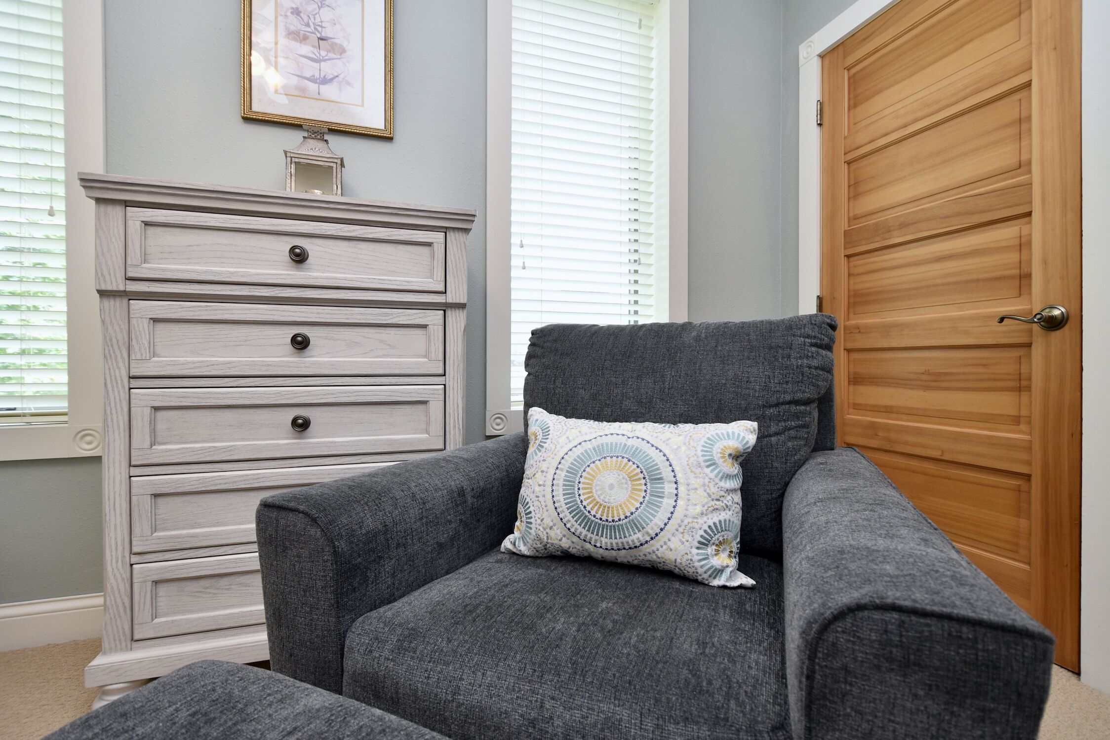 A gray armchair with a patterned pillow, a white dresser with multiple drawers, and a wooden door in a room with light gray walls and a window with blinds.