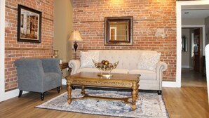 A living room with a brick wall, a white sofa, a blue armchair, a wooden coffee table, and a patterned rug.