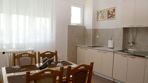 Un coin repas avec une table dressée pour deux personnes, une chaise en bois, un verre à vin et une bouteille de vin. Il y a une fenêtre avec des rideaux légers, un radiateur et un coin cuisine avec un évier et un plan de travail carrelé.