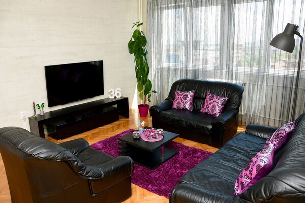 A modern living room with a black leather sofa, a black coffee table, and a TV on a black stand. There is a large potted plant and a purple rug on the floor.