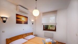 A double bed with a wooden headboard, a wall-mounted air conditioner, a painting of a sunset, a window with blinds, and a bedside lamp. - House Petra on the beach-sea (Cesarica)