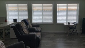 A living room with a sofa, a small table, and a chair. There are three windows with blinds, a heater, and a power outlet on the wall. - Our Cottage on Conesus Lake (Livonia)