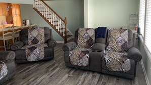 A living room with two grey sofas covered with leopard print throw pillows, a wooden staircase, a kitchen area, and a window with blinds. - Our Cottage on Conesus Lake (Livonia)