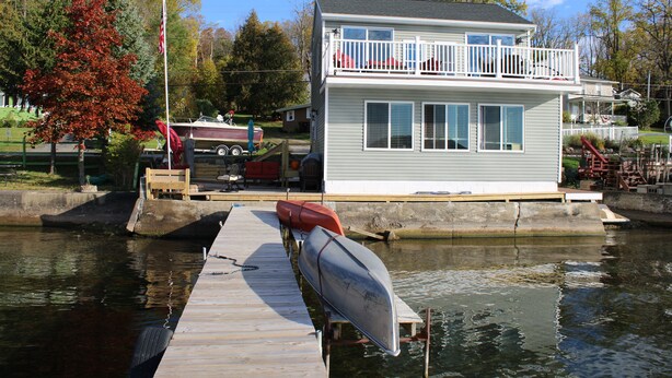 A house with a dock extending into a body of water, a boat on the dock, and a flagpole with a flag. - Our Cottage on Conesus Lake (Livonia)