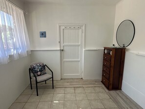 Una habitación con una puerta blanca, un sillón con cojín floral, un aparador de madera con espejo y un cuadro pequeño en la pared.