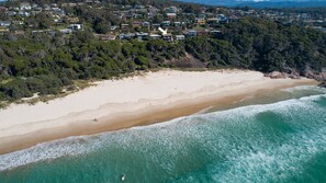 Vista aérea de una zona costera con una playa arenosa, olas oceánicas y una área residencial con casas y árboles.