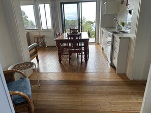 A dining area with a wooden table and chairs, a wicker chair with a cushion, and a view through a glass door to the outdoors.