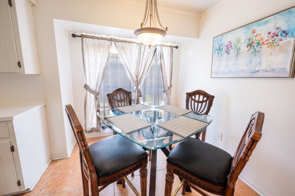 A dining area with a glass-top table, four wooden chairs with cushioned seats, a chandelier, and a painting of flowers on the wall.
