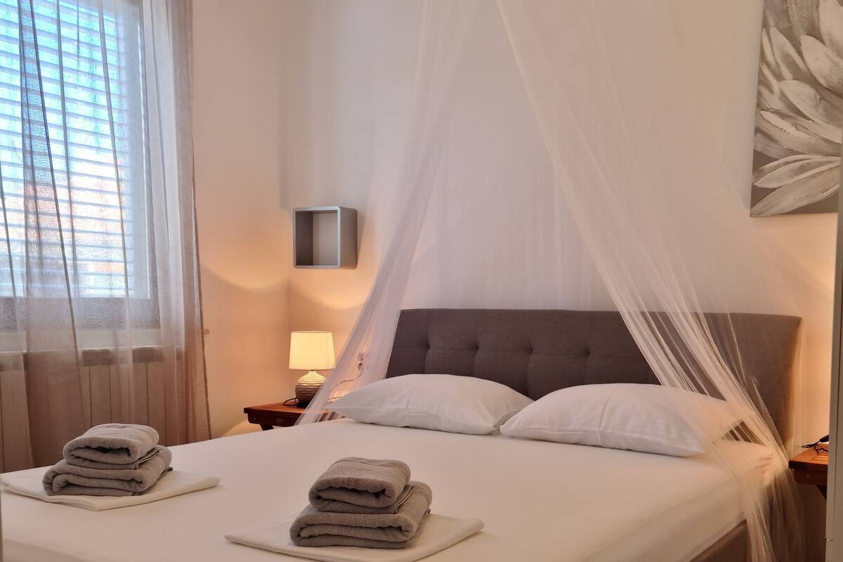 A neatly made bed with a grey headboard, white pillows, and a stack of folded towels on the side.