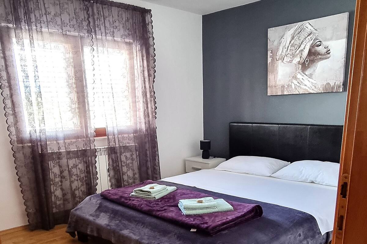 A bedroom with a bed, a nightstand, a lamp, a window with curtains, and a painting on the wall.