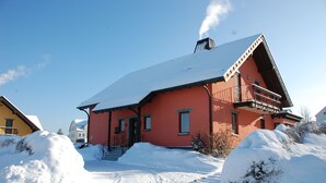 Punainen mökki, jonka lumipeitteinen katto ja savupiipusta nouseva savu erottuvat selvästi, ja sen ympärillä on lunta.