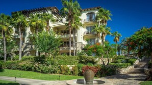 A multi-story hotel with balconies, surrounded by lush greenery and a stone pathway leading to a central fountain.