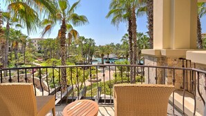 A balcony with wicker furniture overlooking a pool area surrounded by palm trees and a building in the background.