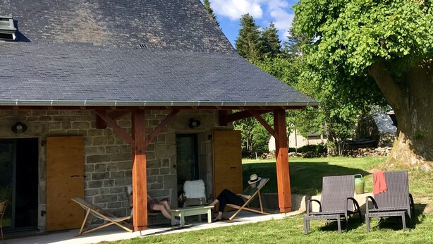 Ein Steinhaus mit dunklem Dach und einer Holzveranda. Auf der Veranda stehen zwei Stühle und ein Tisch.