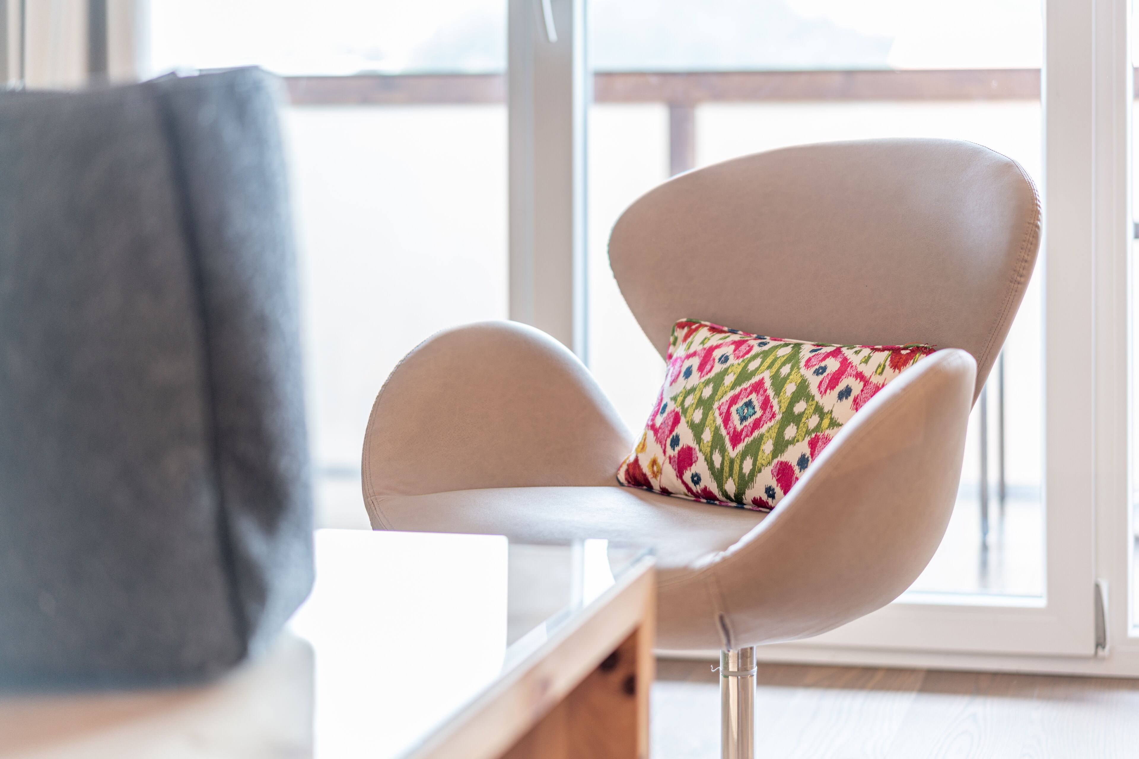 Une chaise moderne avec un coussin coloré, placée dans une pièce lumineuse dotée d’une grande fenêtre.