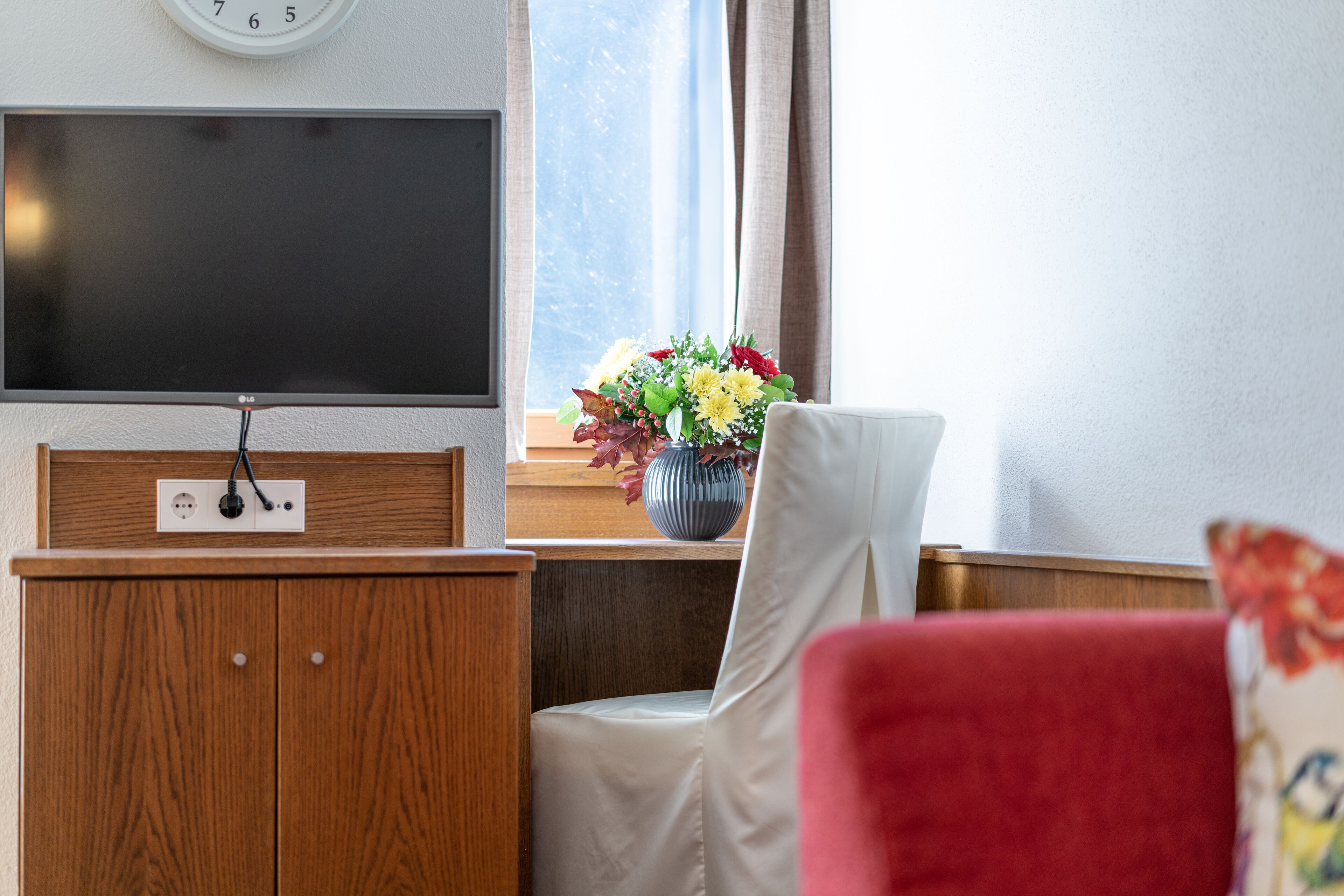 A living room with a flat-screen TV, wooden cabinet, and a window with a flower vase.