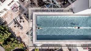 An aerial view of a rooftop swimming pool with two people swimming, surrounded by outdoor seating and a modern architectural design. - Hotel Naudererhof (Nauders)