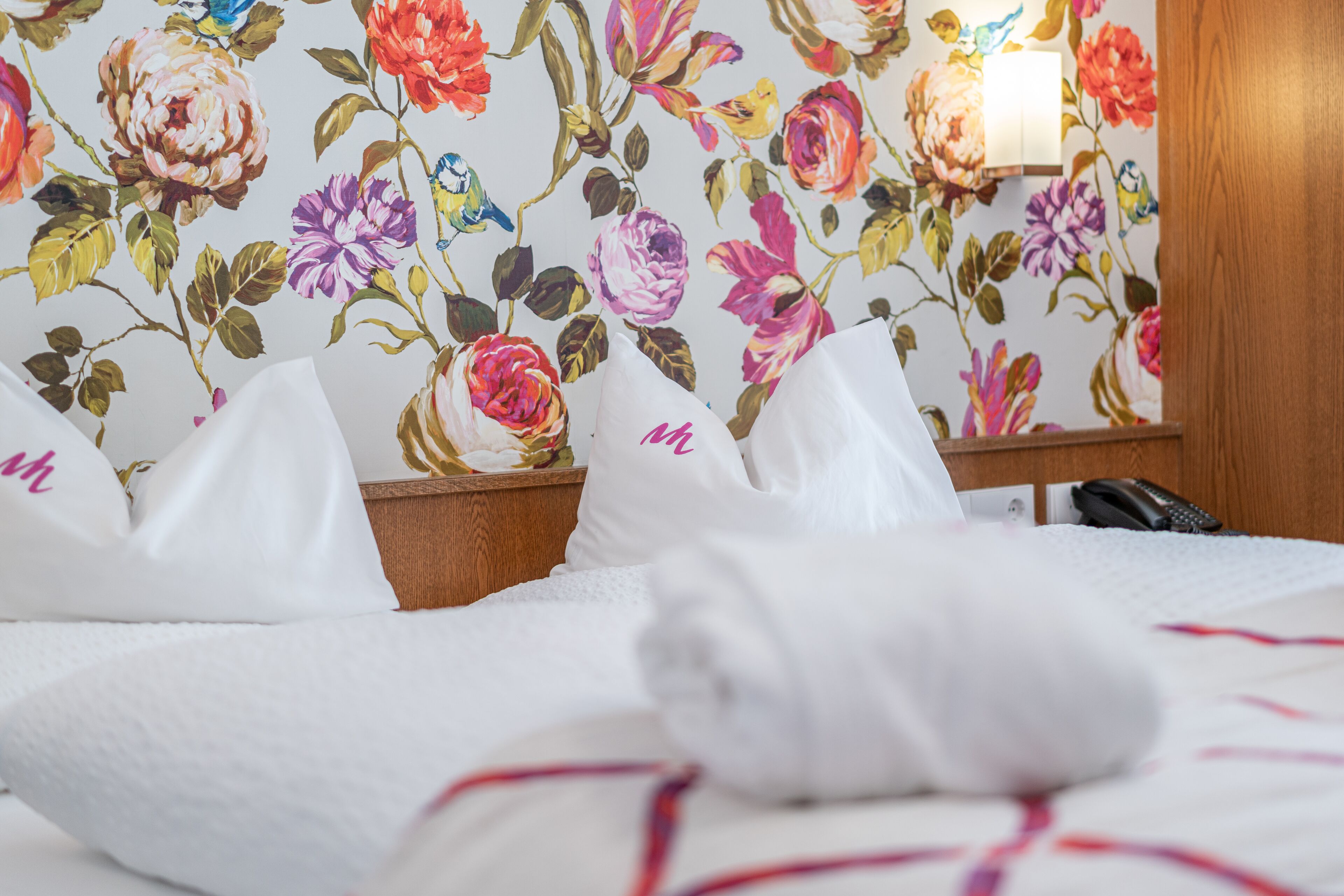 Ein Hotelzimmer mit floraler Tapete, einem ordentlich bezogenen Bett mit weißen Leinen und einer Nachttischlampe.