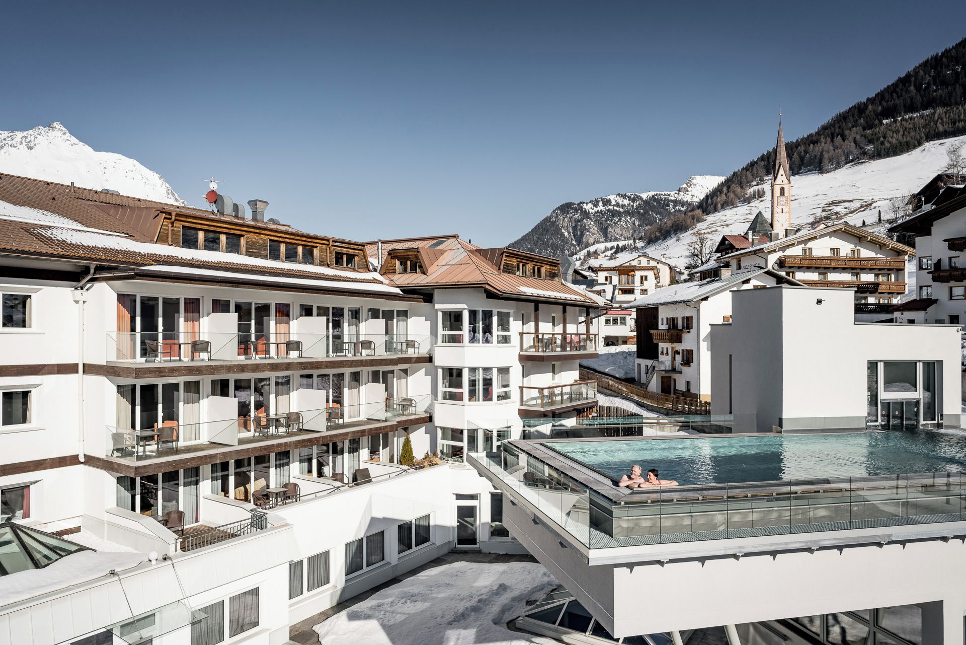A modern hotel with multiple balconies, a swimming pool, and a mountain backdrop.