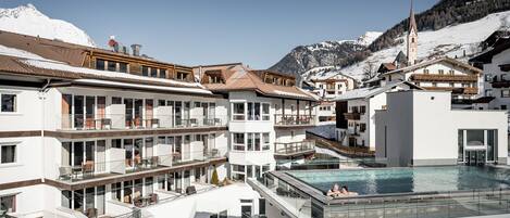 A modern hotel with multiple balconies, a swimming pool, and a mountain backdrop.