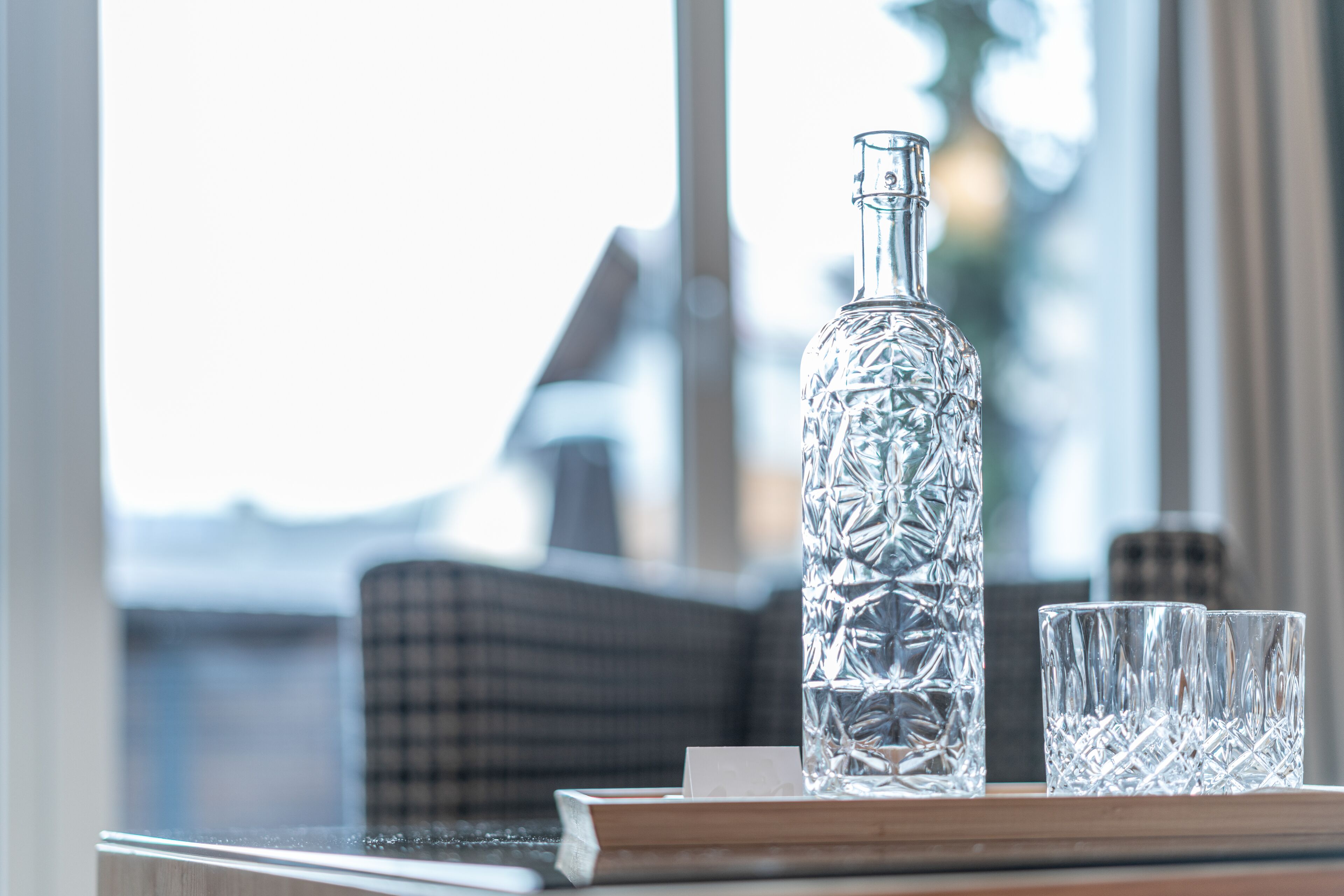A clear glass bottle and two glasses on a table with a checkered cushion in the background.