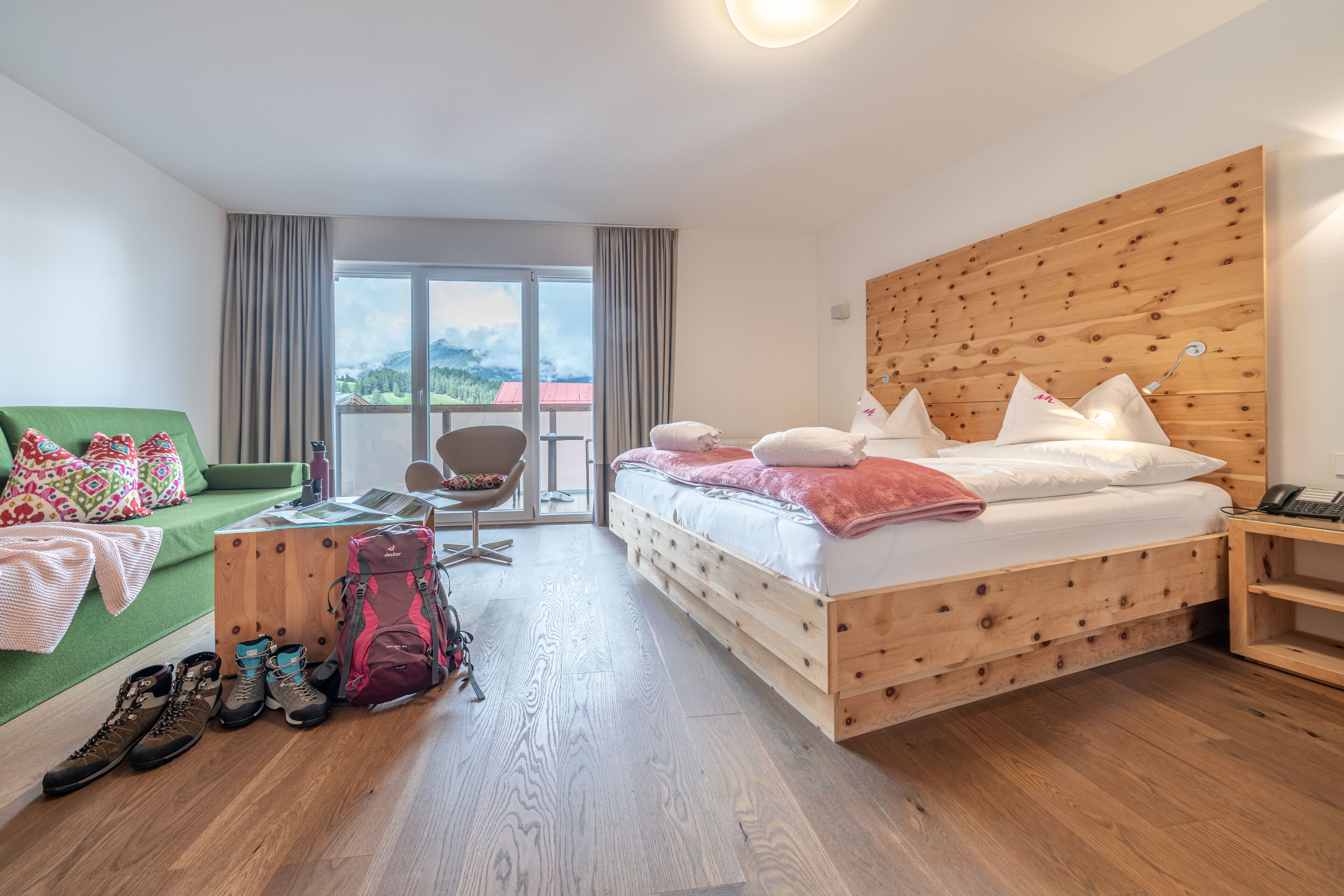 Ein Hotelzimmer mit einem hölzernen Bett, einem grünen Sofa, einem Sessel und Blick auf die Berge.