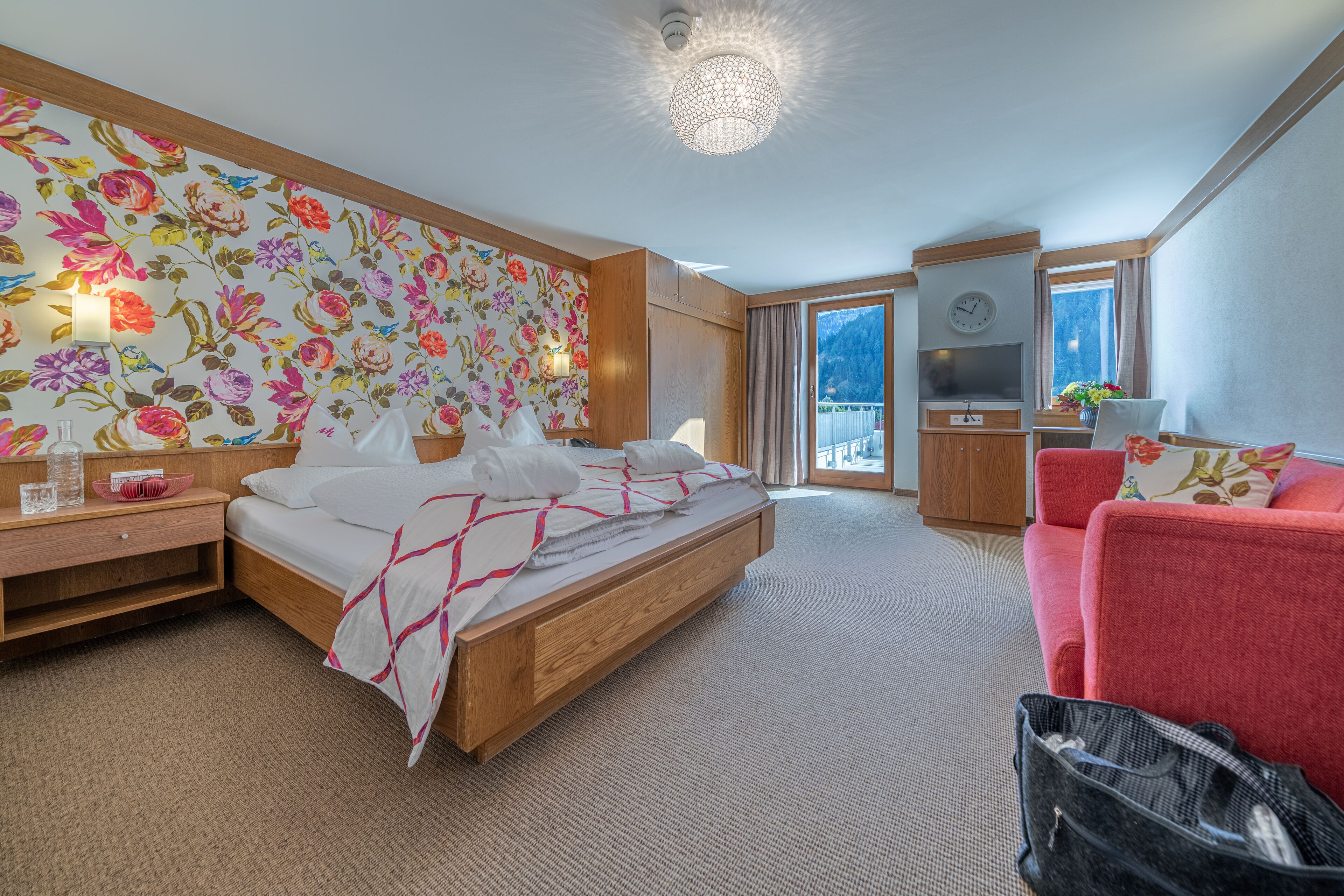 Habitación de hotel con una cama grande, un sofá rojo, un armario de madera, un televisor y un balcón con vistas a la montaña.