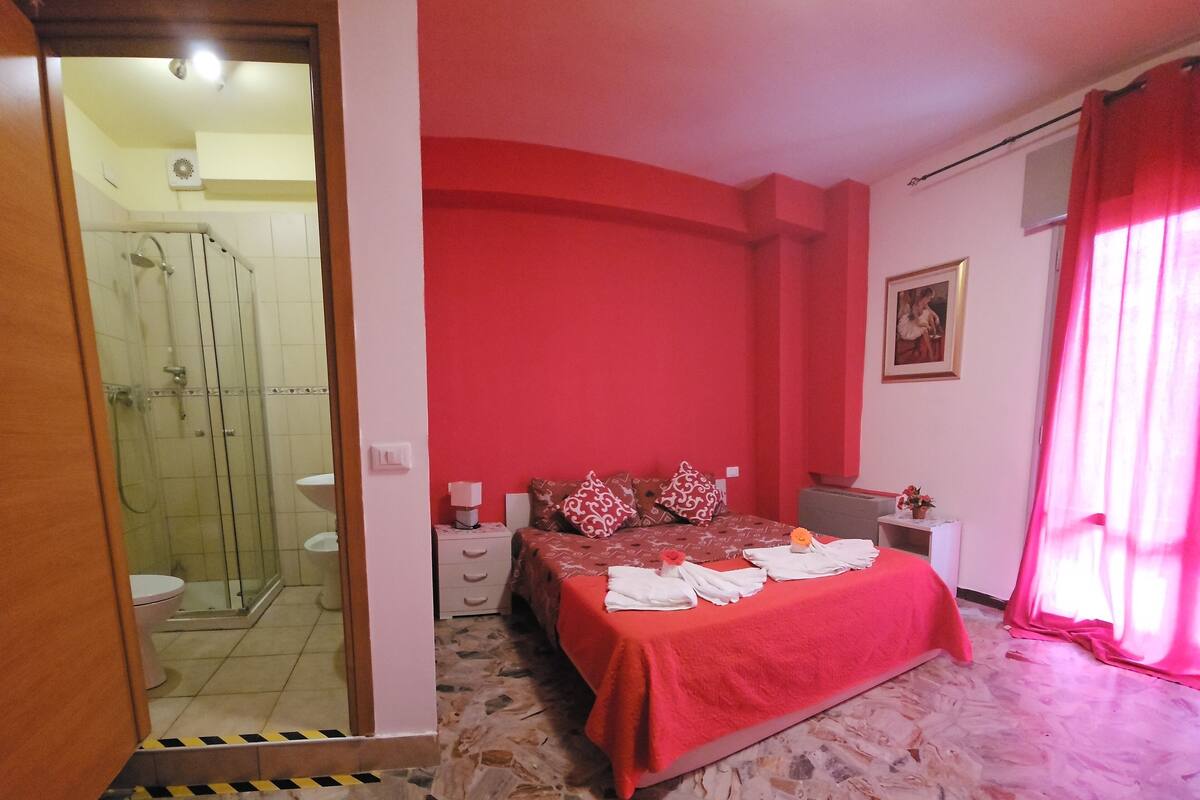 A bedroom with a red bedspread, a shower, a toilet, and a small table with a plant.