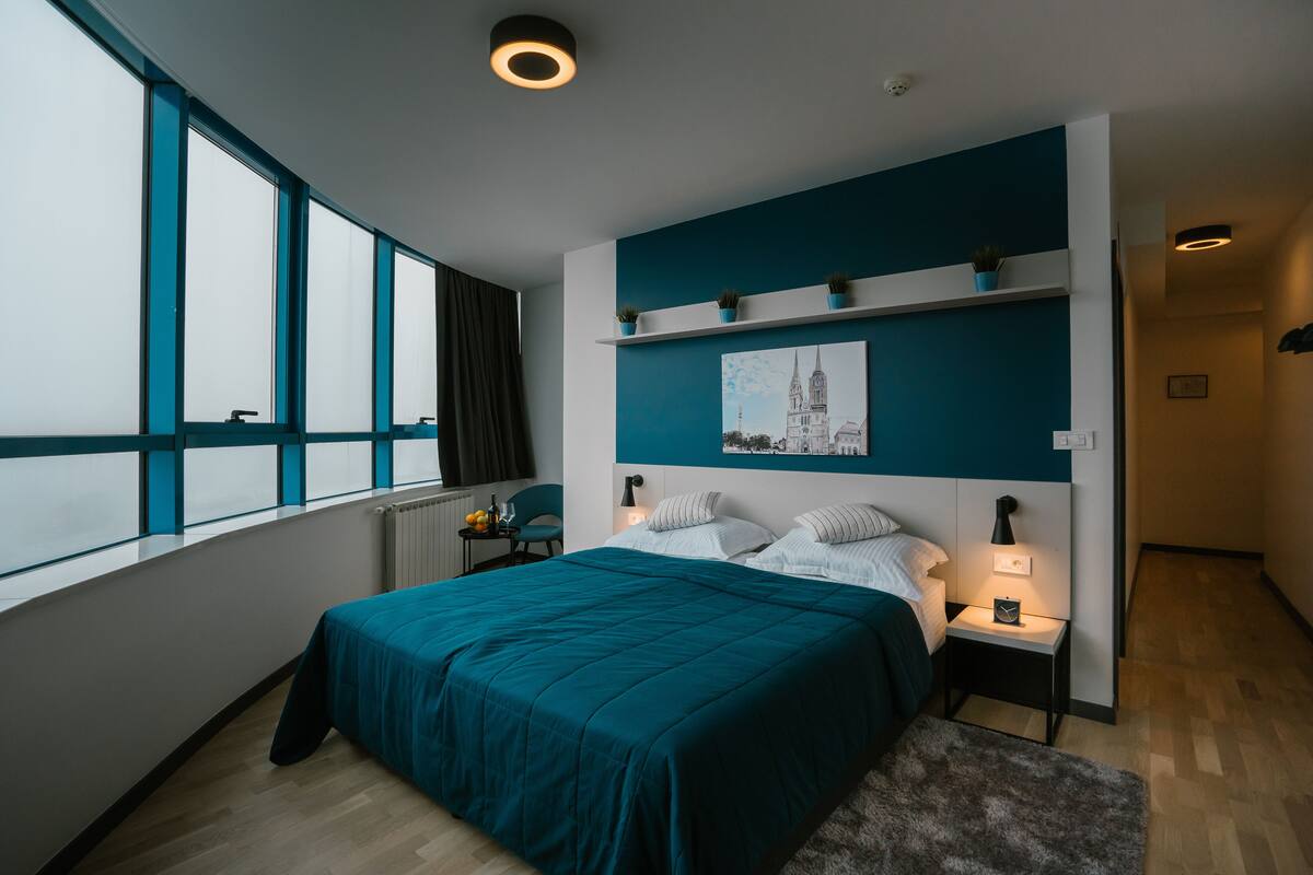 A modern bedroom with a large bed, bedside tables, a nightstand, a window with curtains, a wall-mounted shelf with plants, and a framed picture.