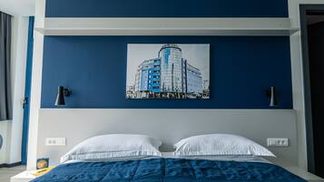 A bedroom with a bed, two white pillows, a blue blanket, a wall-mounted lamp, and a cityscape painting.