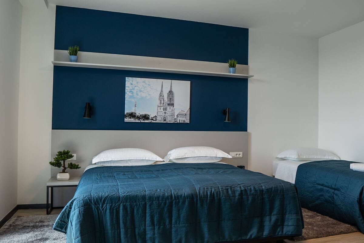 A bedroom with two beds, a nightstand with a plant, a wall-mounted shelf with two potted plants, and a framed picture of a building.