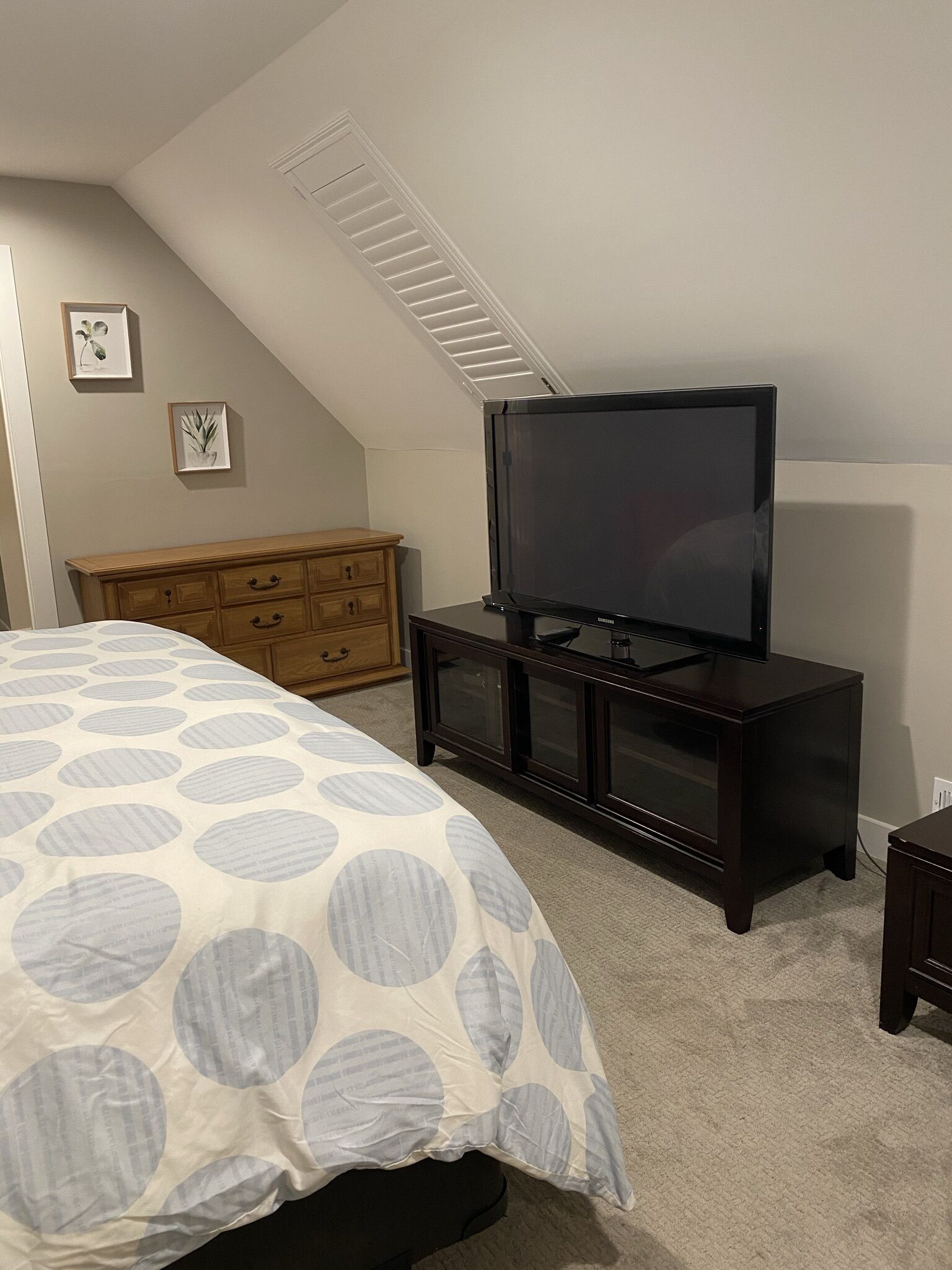 A bedroom with a bed, a TV on a dark wooden stand, a wooden dresser, and a sloped ceiling with a vent.