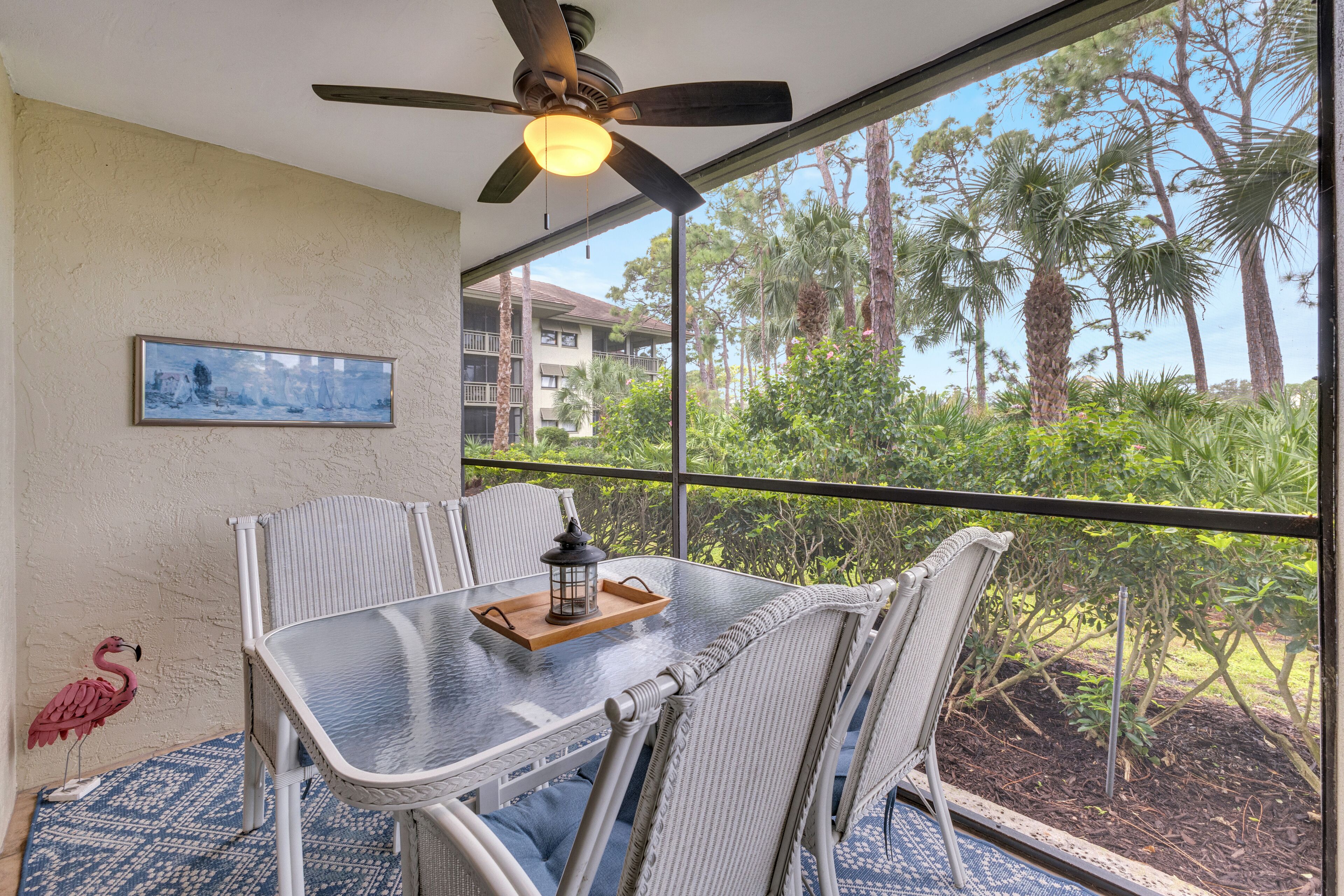 Un coin repas avec une table en verre, des chaises en osier, un ventilateur de plafond et une vue sur des palmiers et un immeuble.