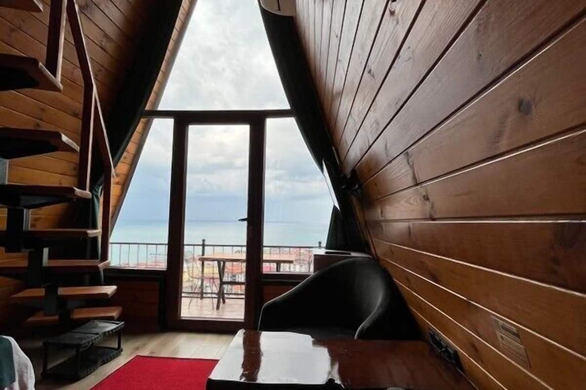 Een knus interieur met houten details, een schuine kap, een rood tapijt en uitzicht op het strand door een groot raam.