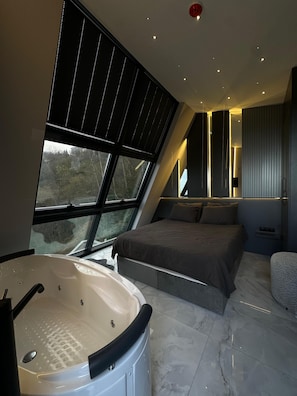 A modern bedroom with a large bed, a bathtub, and a view through a window.