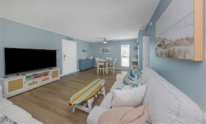 A living room with a flat-screen TV, a sofa, a bench, a dining table with chairs, and a kitchen area.
