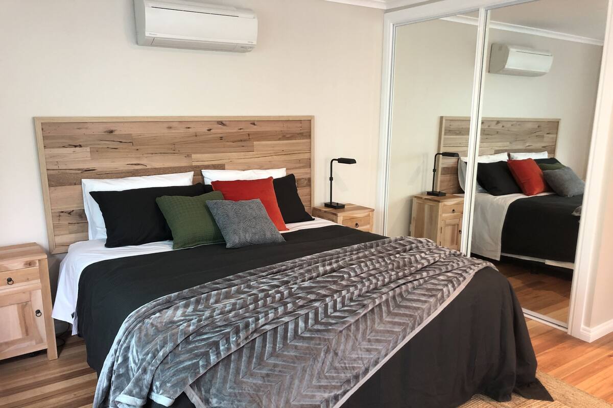 A neatly arranged bedroom with a wooden headboard, a large bed with multiple pillows, a bedside table with a lamp, and a mirror reflecting the room.