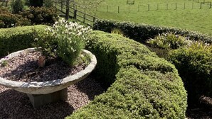 Un jardín ajardinado con un parterre de piedra circular, setos cuidadosamente recortados y vistas a un campo verde con árboles y montañas a la distancia.