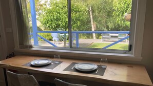 Una mesa de comedor con dos sillas, una ventana que ofrece vistas a follaje y una estructura azul al exterior.