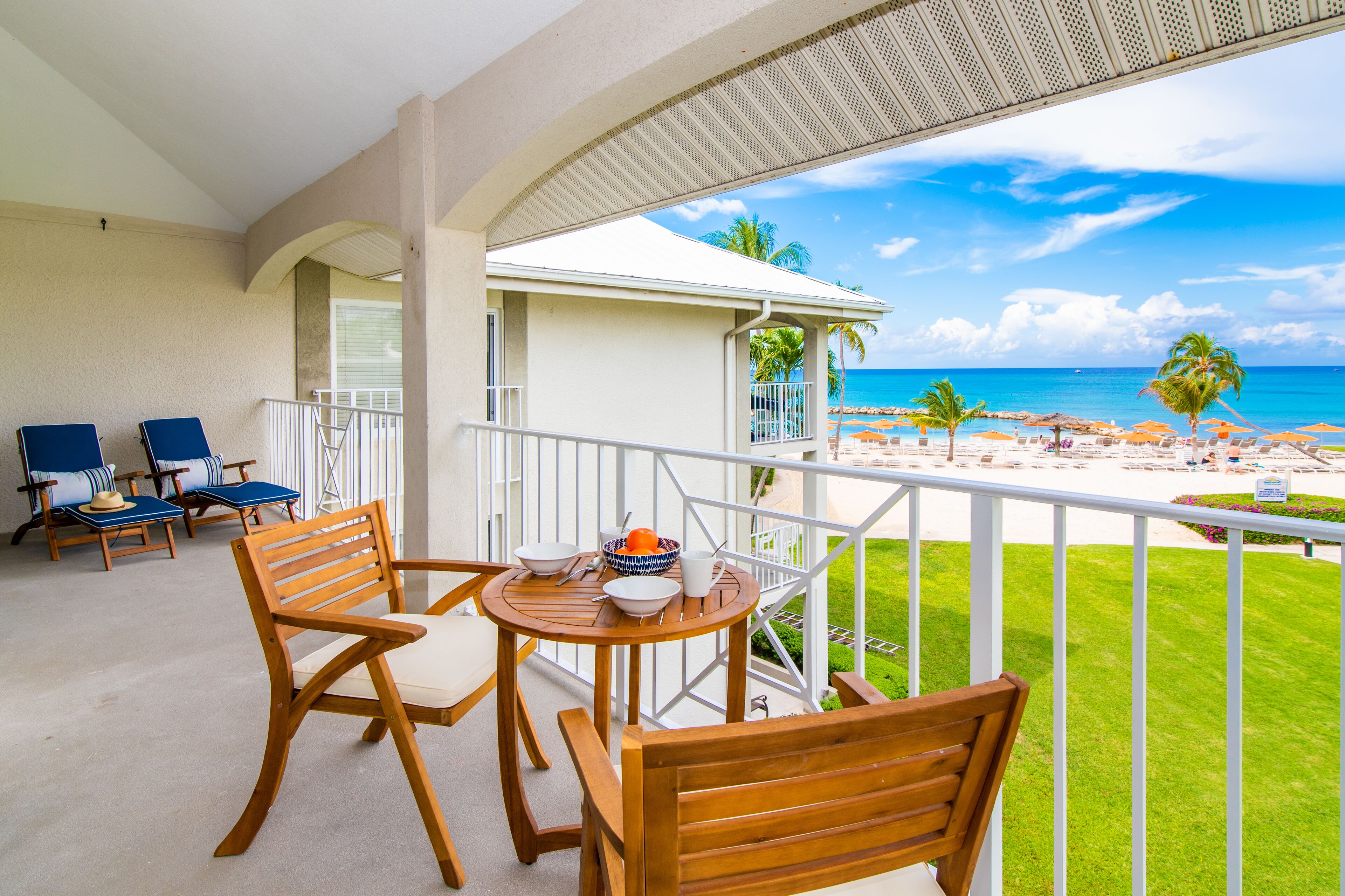 Un balcon avec des chaises et une table en bois, donnant sur une plage bordée de palmiers et surplombant un ciel bleu azur.
