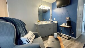 Ein modernes Wohnzimmer mit einem grauen Sofa, einem Couchtisch, einem Laptop, einem an der Wand montierten Fernseher und einem Badezimmer mit Waschbecken und Spiegel.